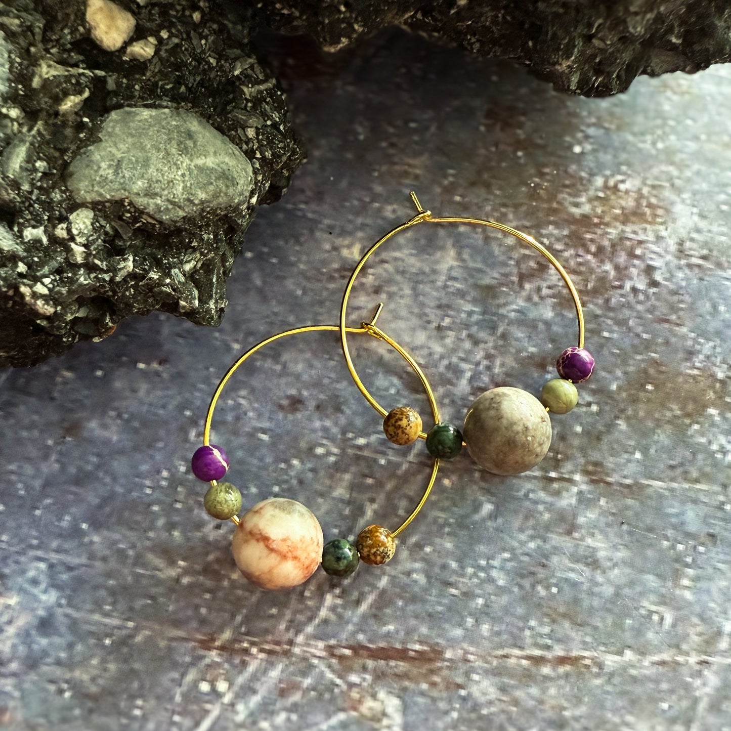 Gold hoop earrings with natural stone beads, displayed on a textured blue/purple surface next to a dark rock.