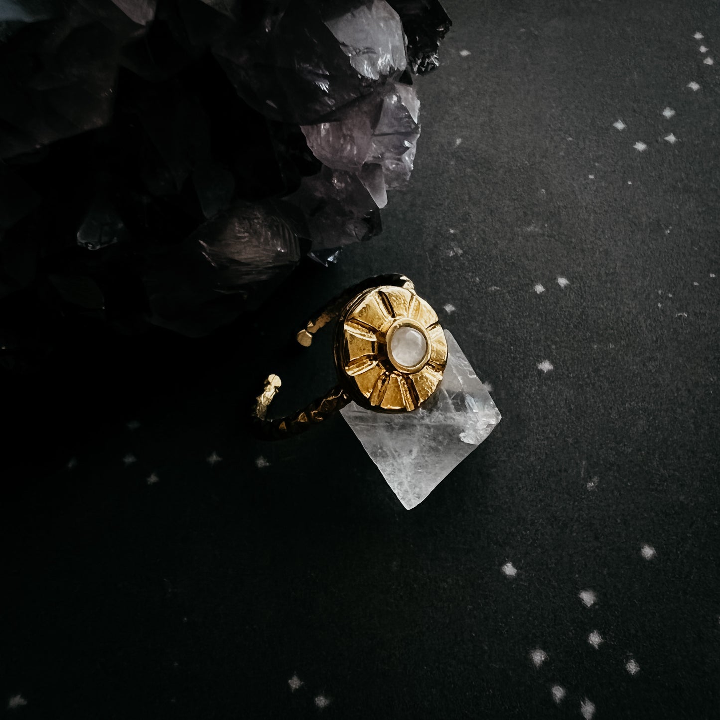 A gold ring with a sunburst shaped setting and a moonstone center, resting on a clear crystal against a dark textured background with black crystals.