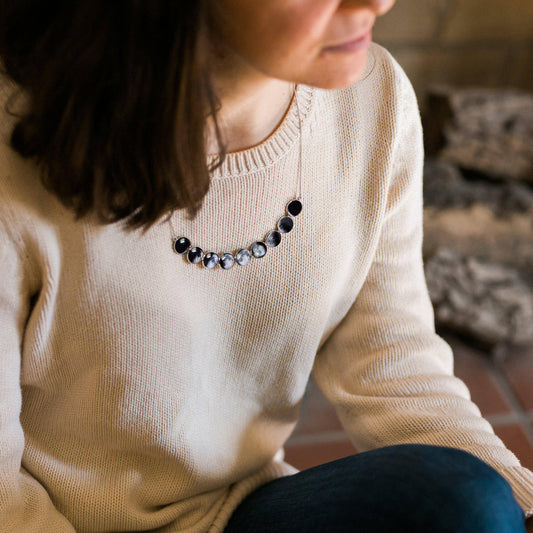 Person wearing a beige sweater with a moon phase necklace, sitting indoors.