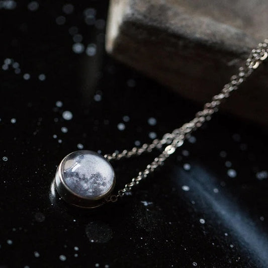 A silver-toned chain necklace with a round pendant featuring a moon design inside a clear dome, displayed on a dark, starry surface next to a stone.