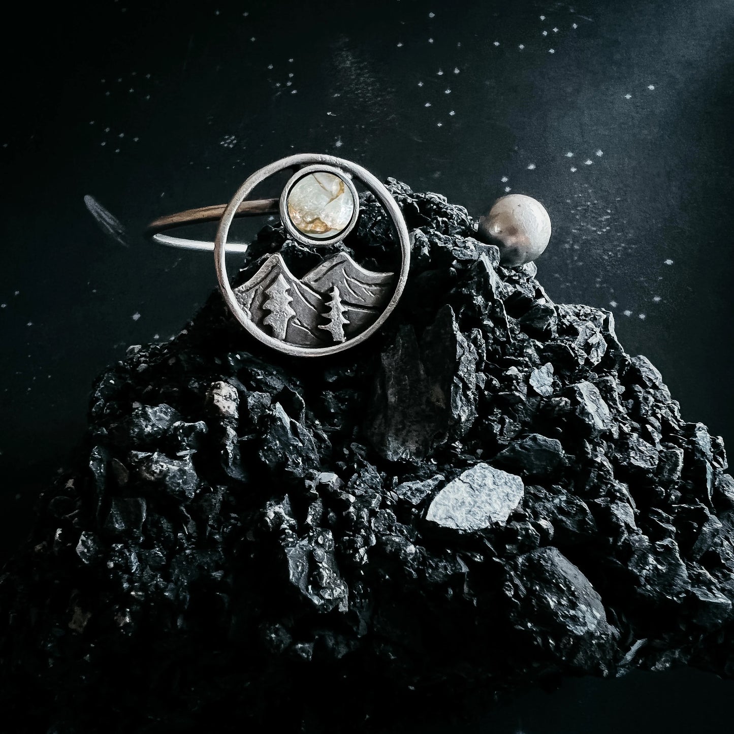 A silver-toned ring with a circular mountain and tree motif and a small oval stone, displayed on a dark textured rock against a starry black background.