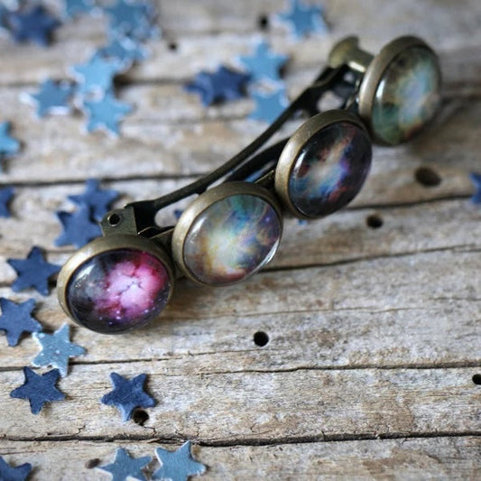 A small barrette with galaxy images set in its circles, resting on a rustic wooden surface scattered with blue star-shaped confetti.