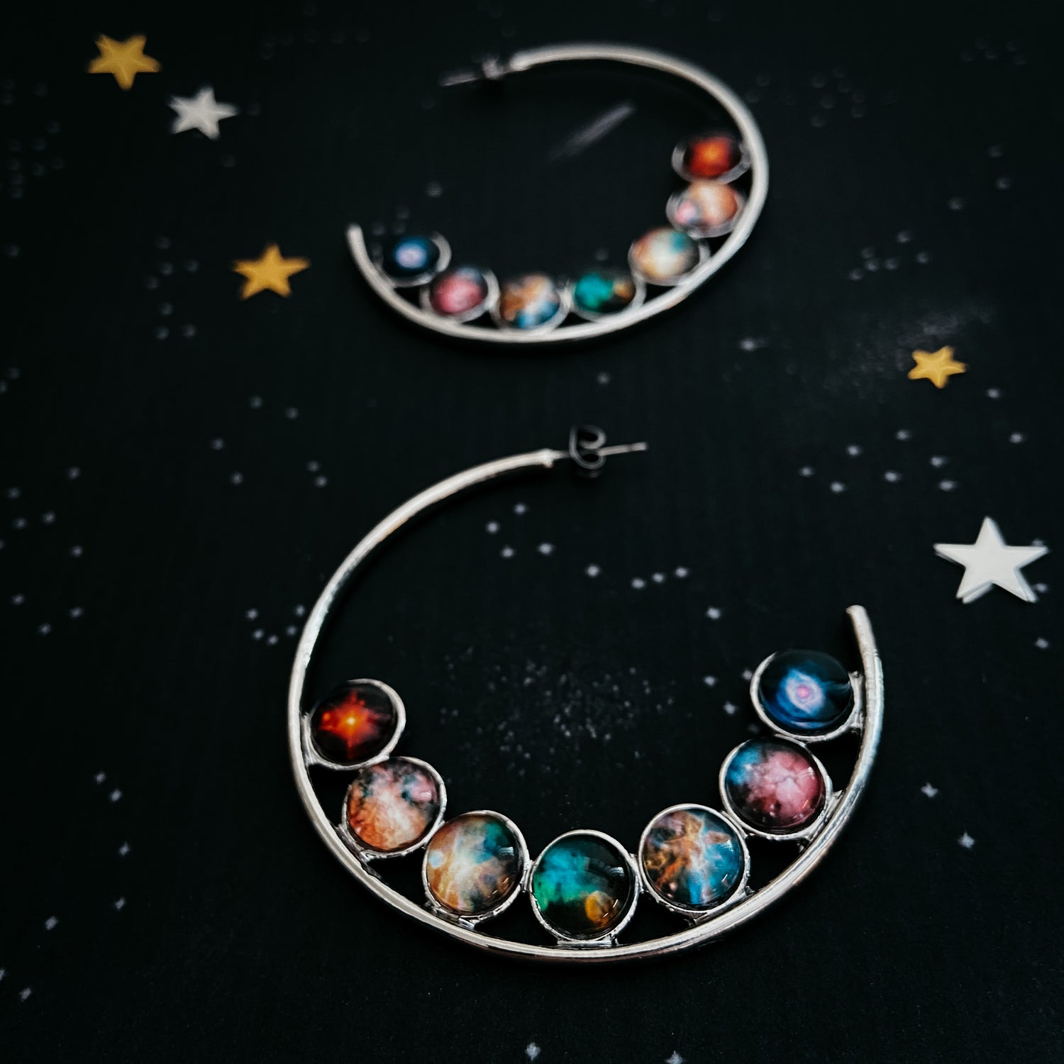 Silver hoop earrings with ROYGBIV images of nebula set in glass. Earrings are laid against a black background with stars. Available at yugenhandmade.com