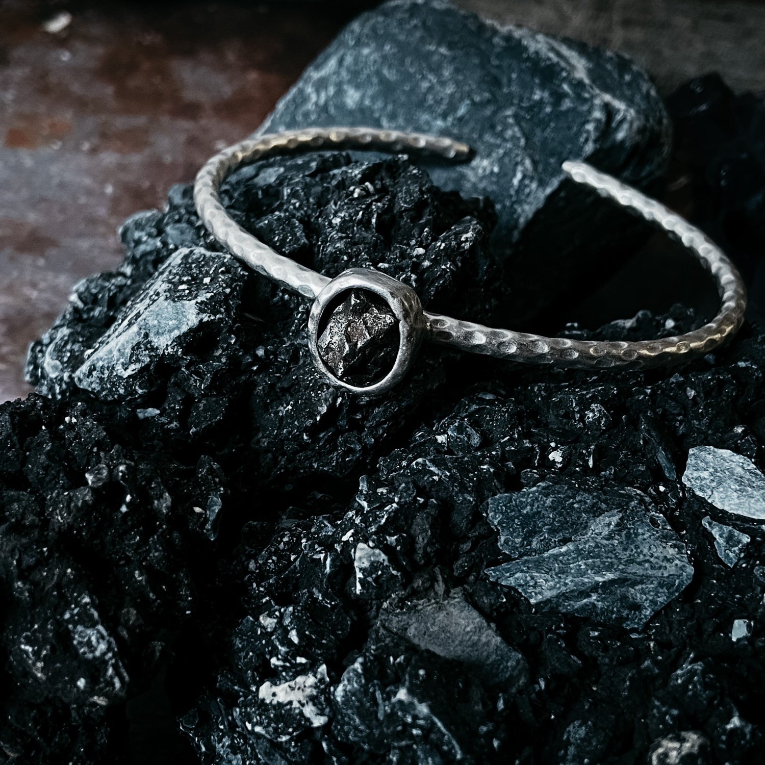 Silver bracelet with hammered texture and oval bezel set with raw campo del cielo meteorite, set on top of a black rock, designed and handcrafted by Yugen Handmade