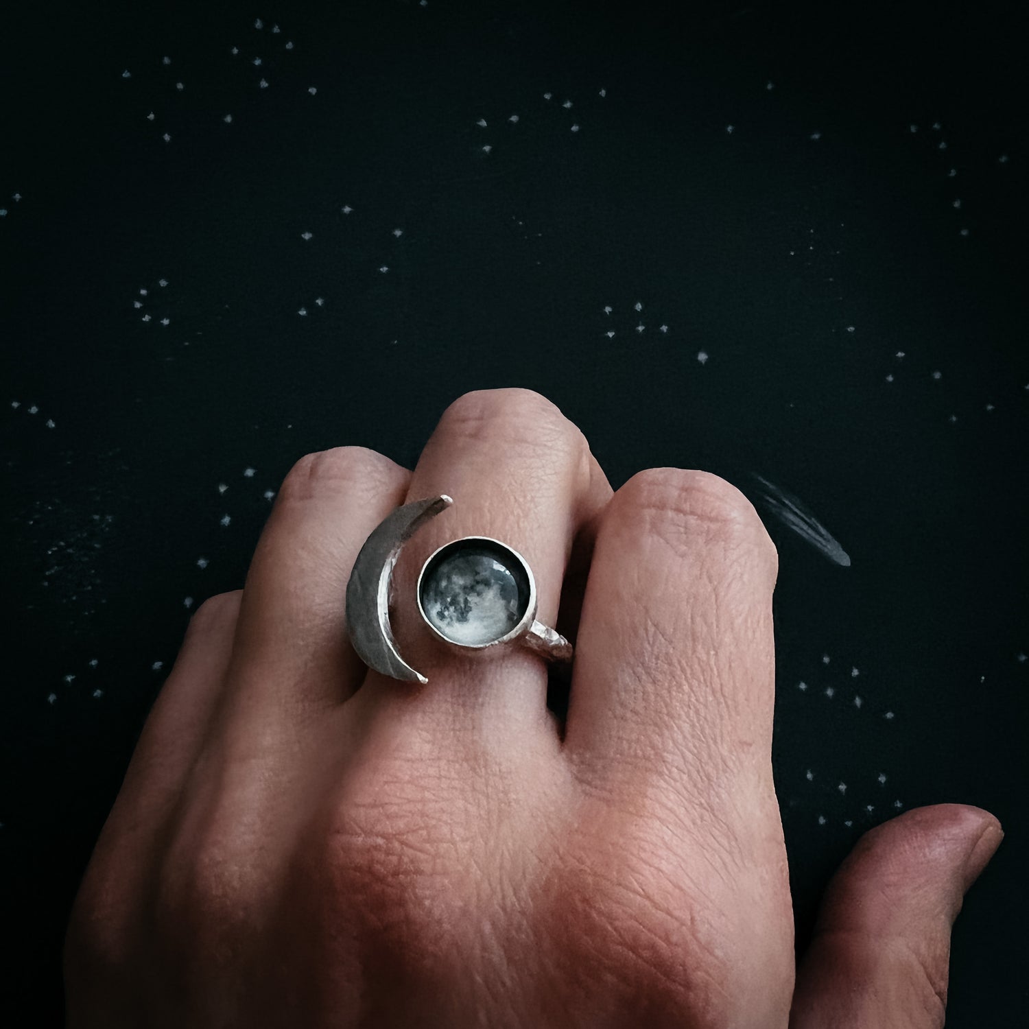 A Caucasian female hand wearing a silver tone ring set with a glass image of the full moon that is flanked by a silver Crescent Moon. Hand is against a black background with stars. Ring designed and handcrafted by Yugen Handmade