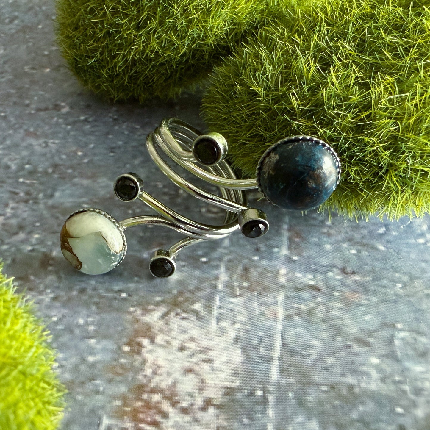 A silver-toned ring with organic-shaped blue and white marbled stones, and small black round stones, displayed on a textured gray surface with green moss props.