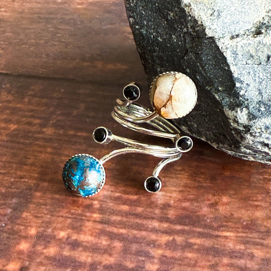 A silver-toned multi-band ring featuring a large round turquoise-blue stone and beige stone, and several small black round stones, displayed on a wooden surface next to a dark rock.