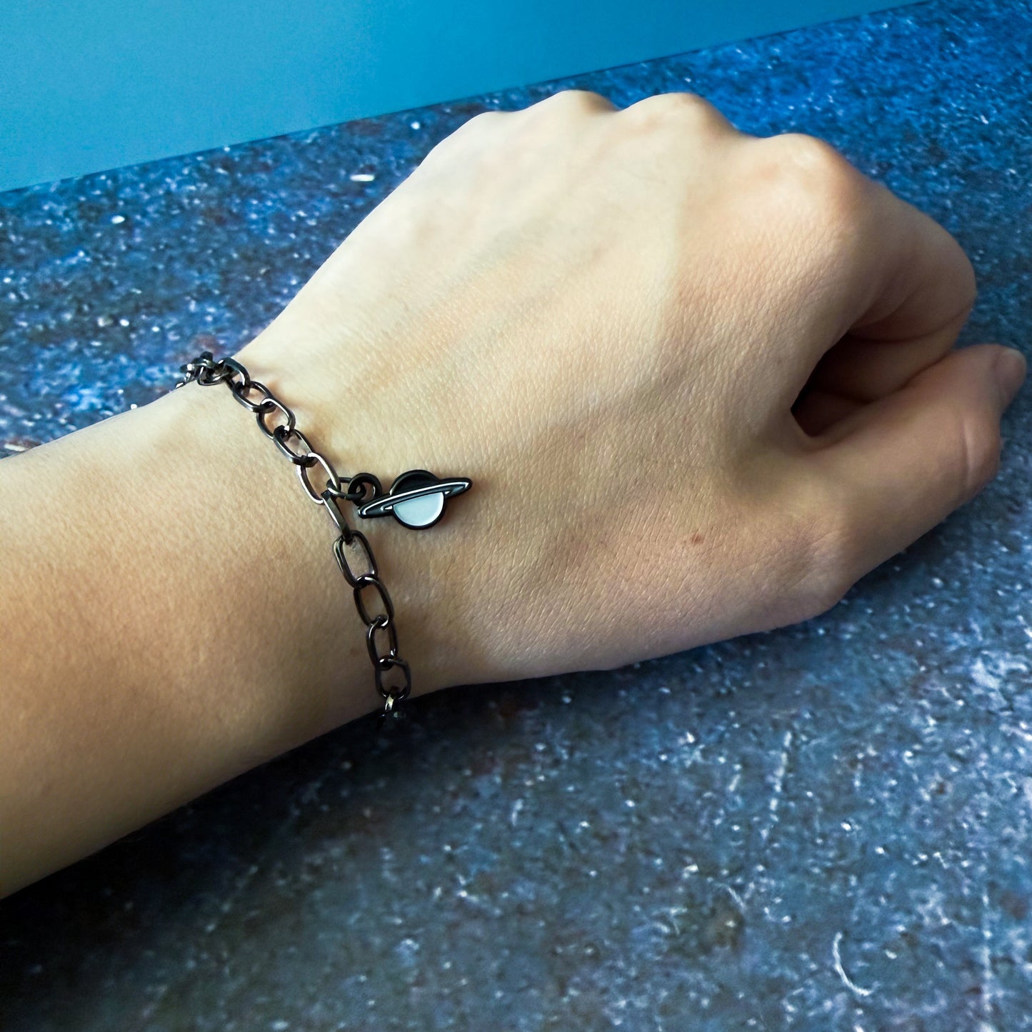 Hand wearing a chain bracelet with a small uranus charm on a blue surface