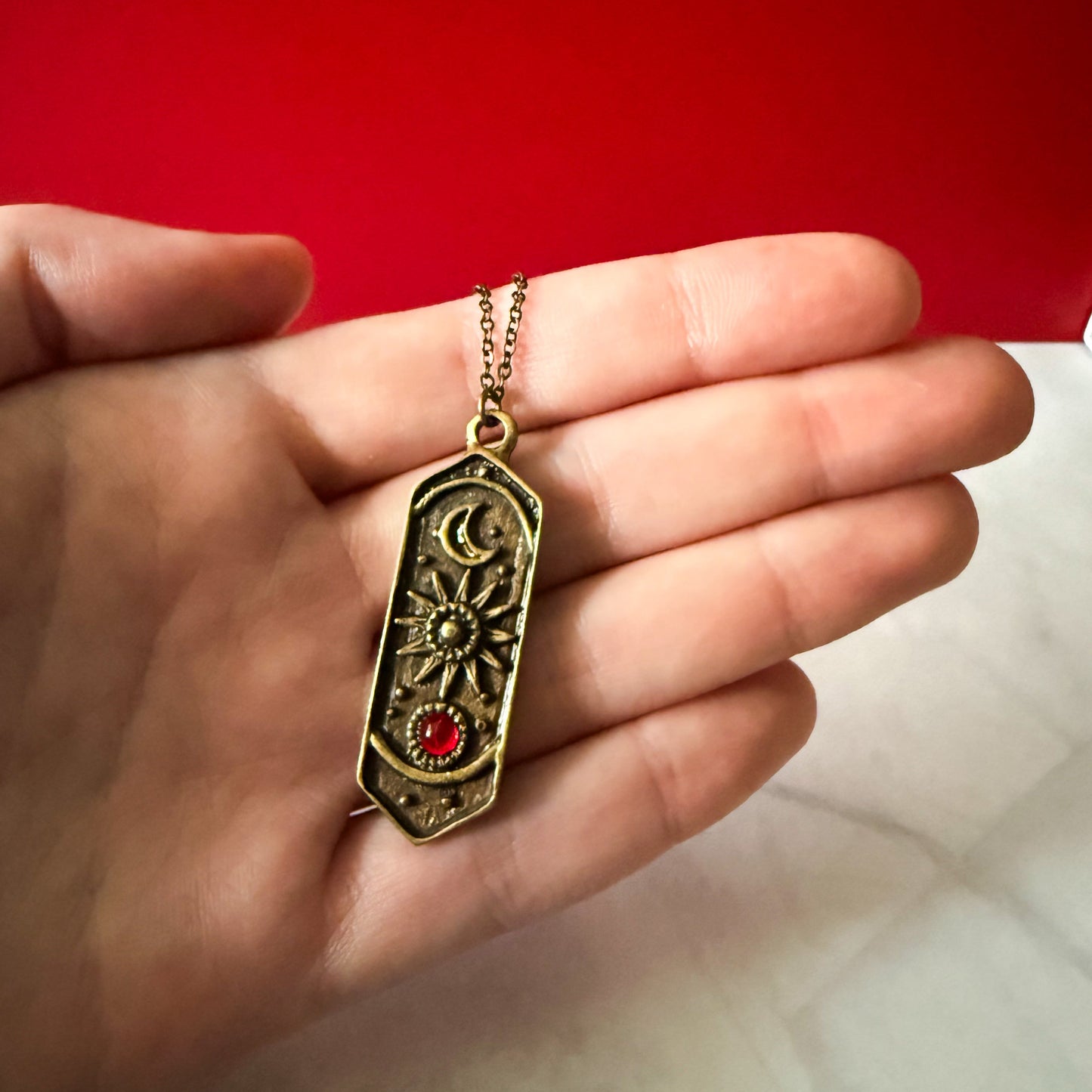 Hand holding a bronze necklace with a decorative pendant against a red background