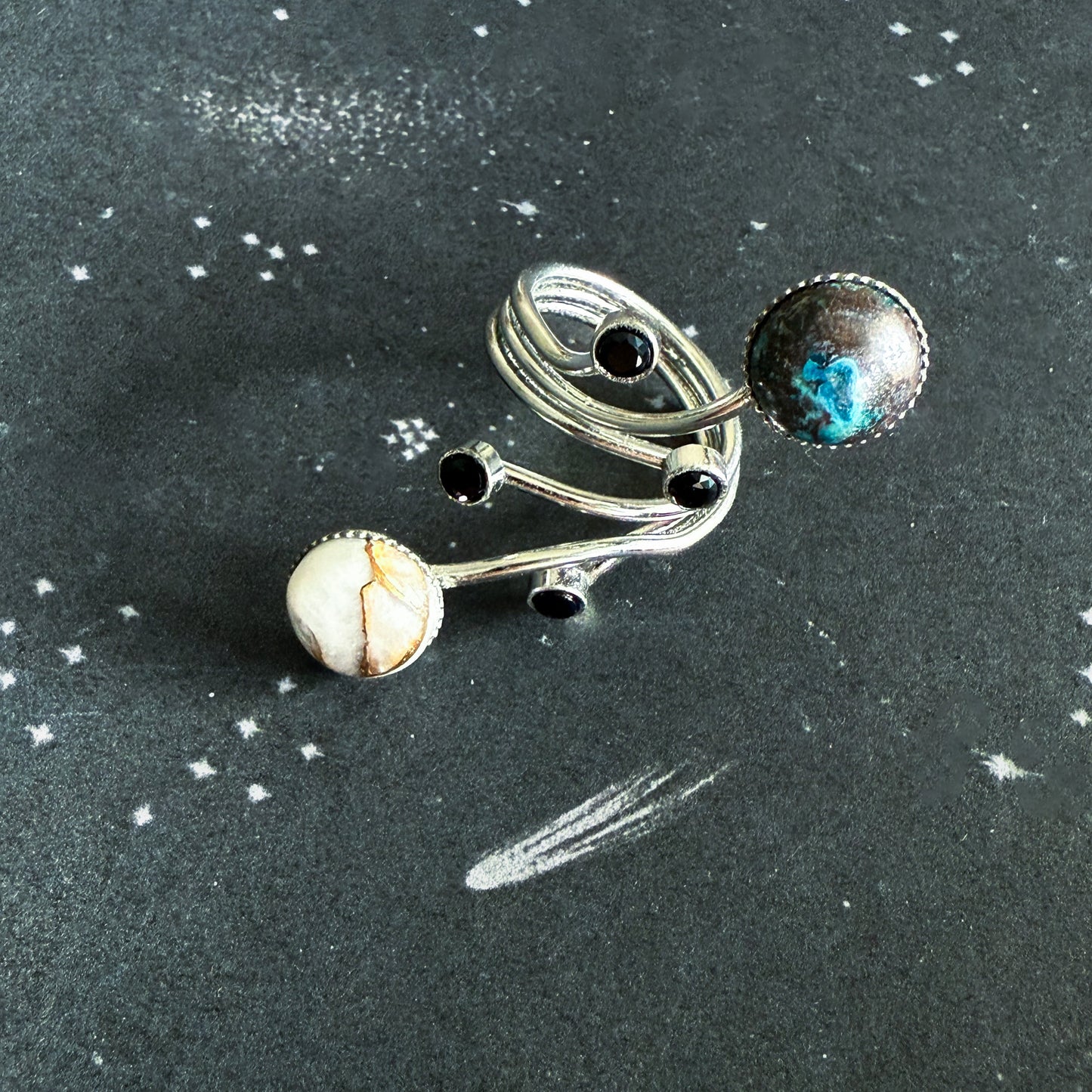 A silver-toned ring with multiple curved bands, small black round stones, and two larger stones—one white with brown veining and one blue-black marbled—displayed on a dark textured background with white speckles.