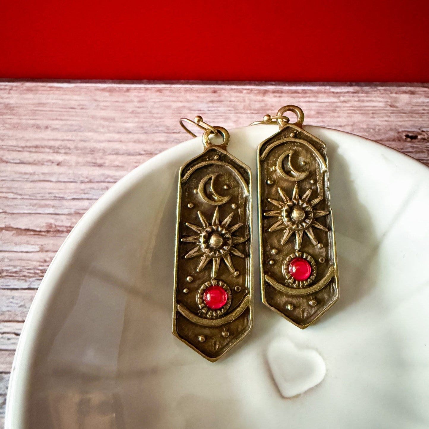 Pair of ornate earrings with birthstones on a white dish against a red background
