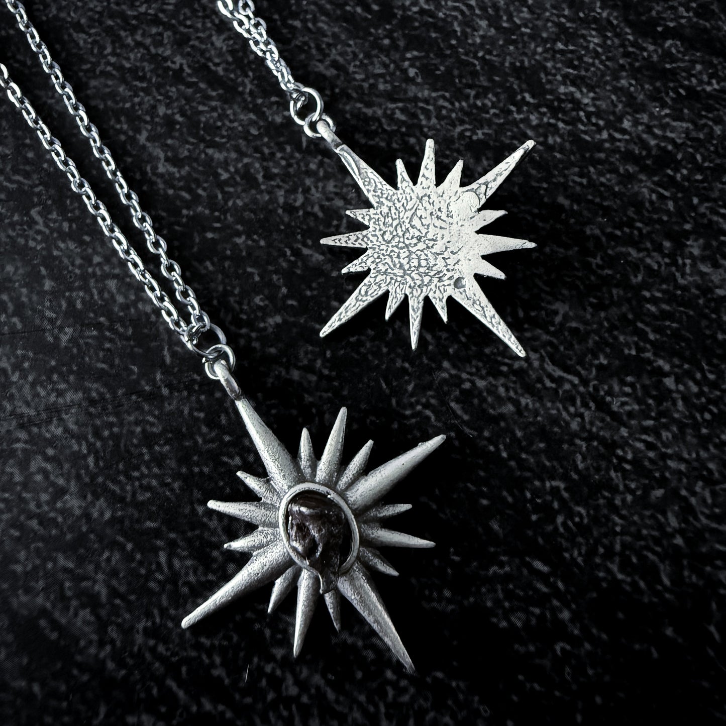 Two sun-shaped silver pendants on a chain against a dark background
