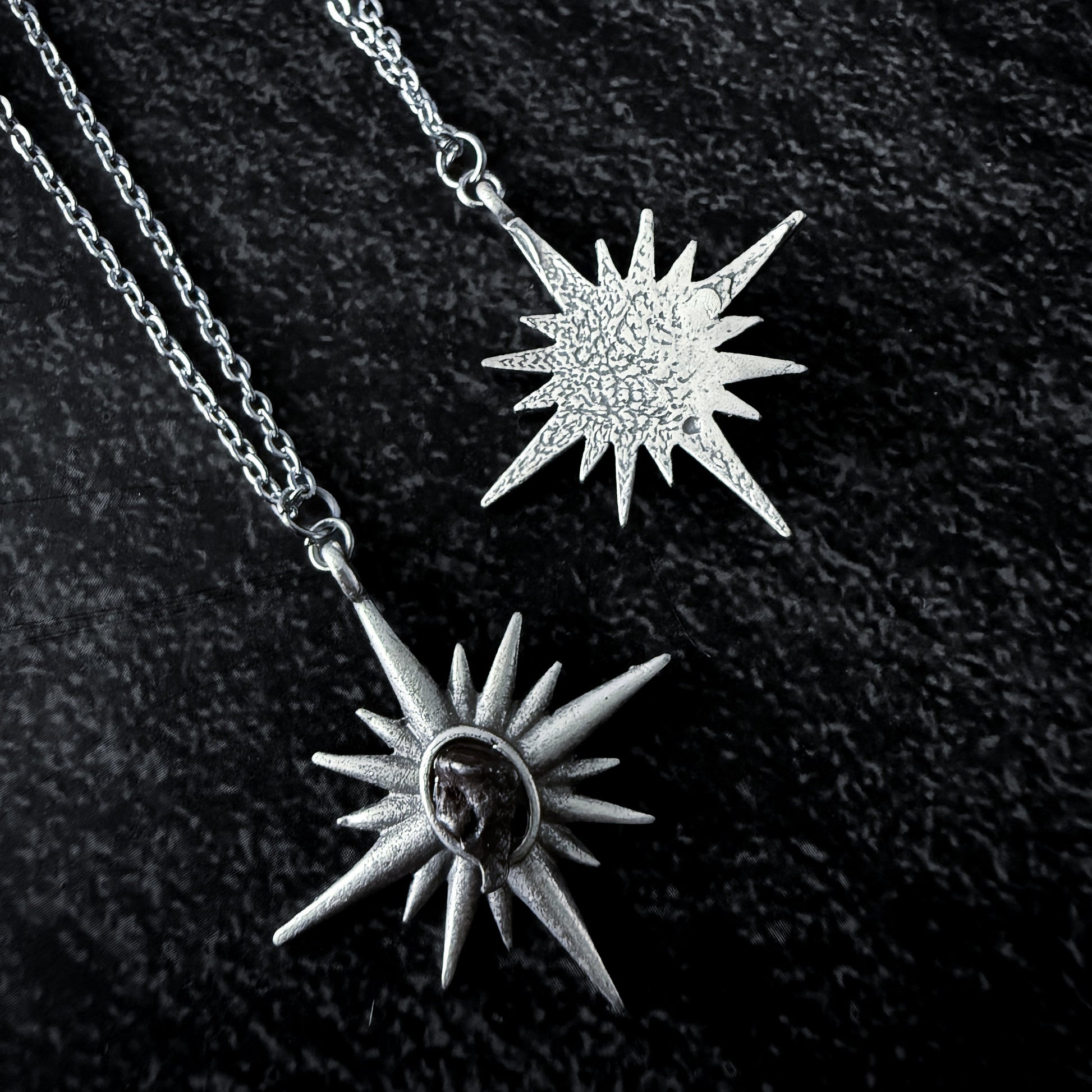 Two sun-shaped silver pendants on a chain against a dark background