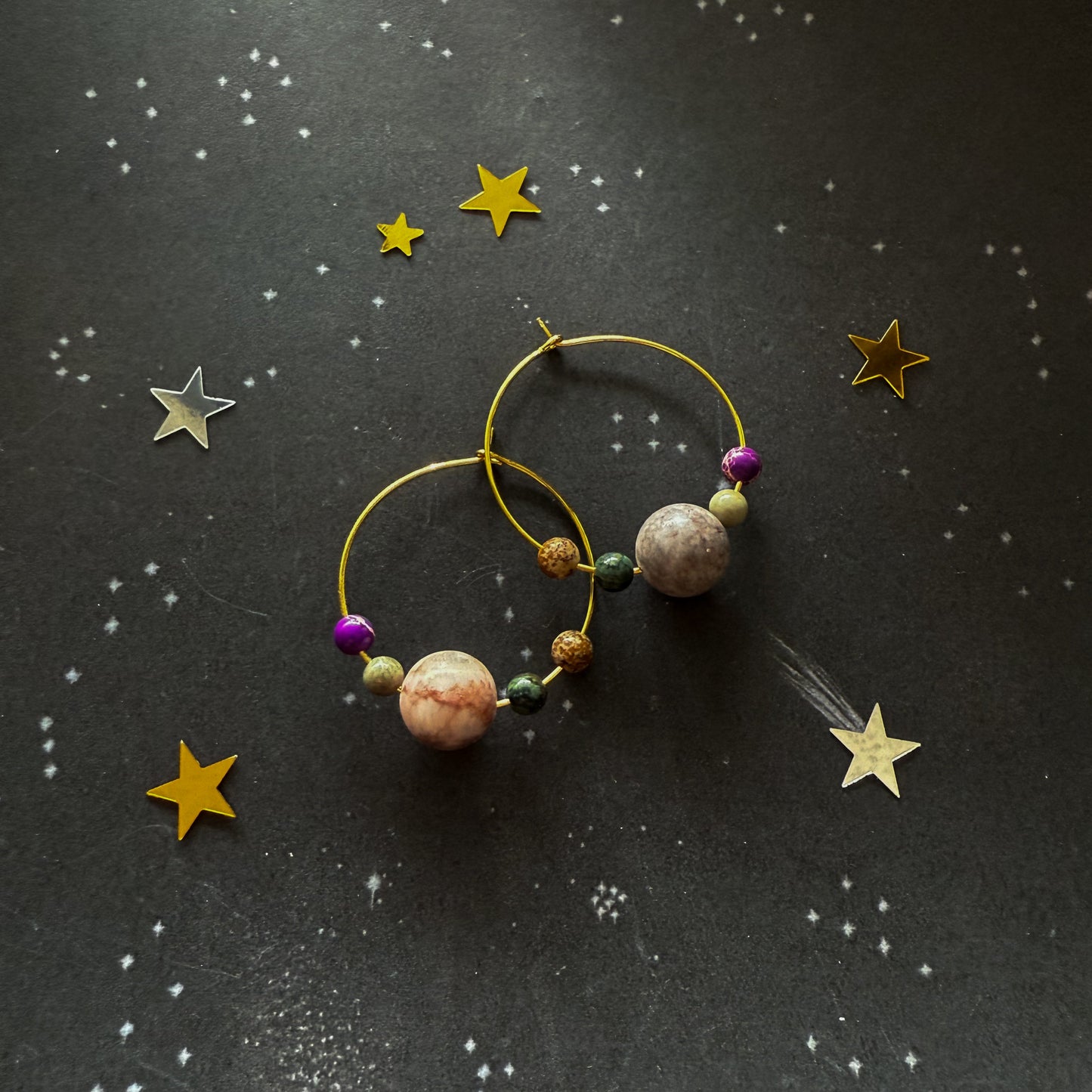 Gold hoop earrings with large marbled beads and small colorful spacer beads, arranged on a dark starry background.
