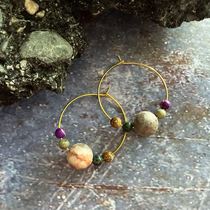 Gold hoop earrings with natural stone beads, displayed on a textured blue/purple surface next to a dark rock.