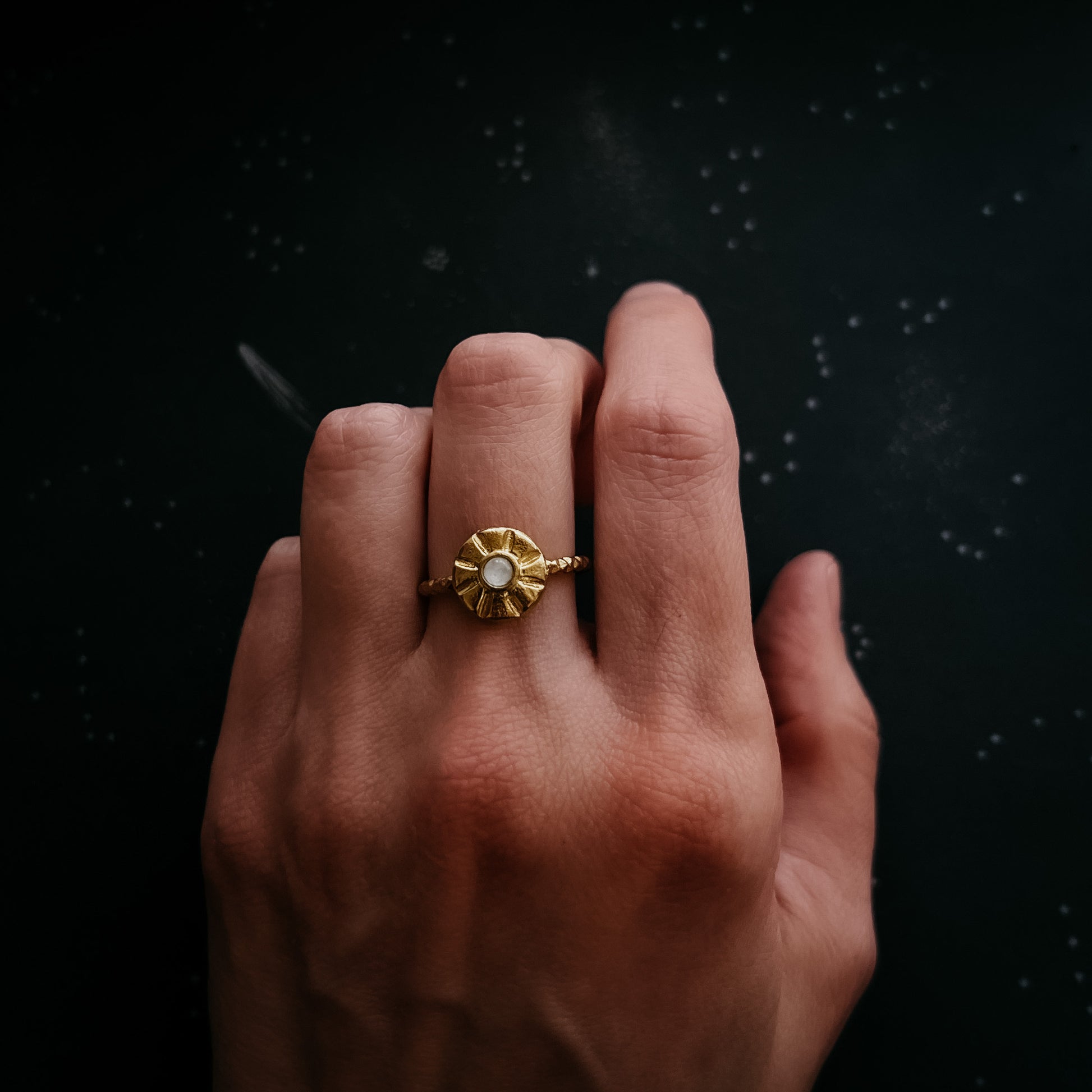 A gold ring with a textured, sun burst circular setting and a central moonstone, worn on a person's finger against a dark, speckled background.