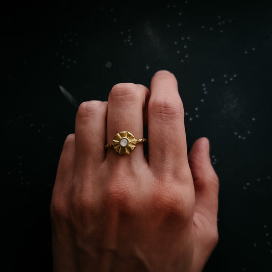 A gold ring with a textured, sun burst circular setting and a central moonstone, worn on a person's finger against a dark, speckled background.