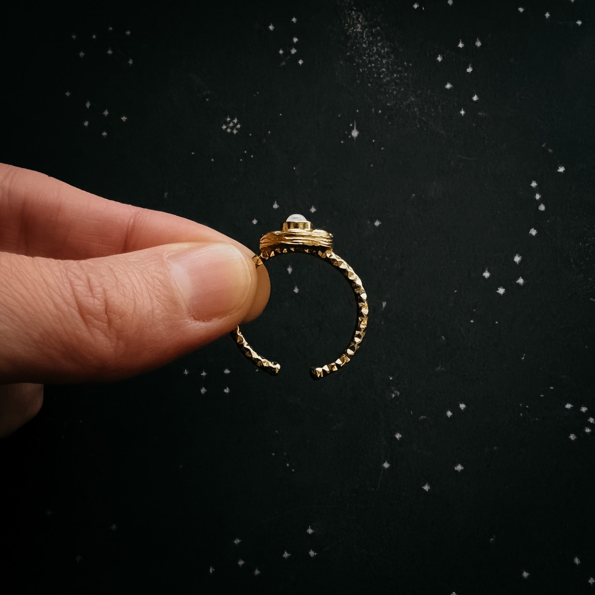 A gold-toned open ring with a textured band and a small dome-cut moonstone on the top, held between two fingers against a dark starry background.