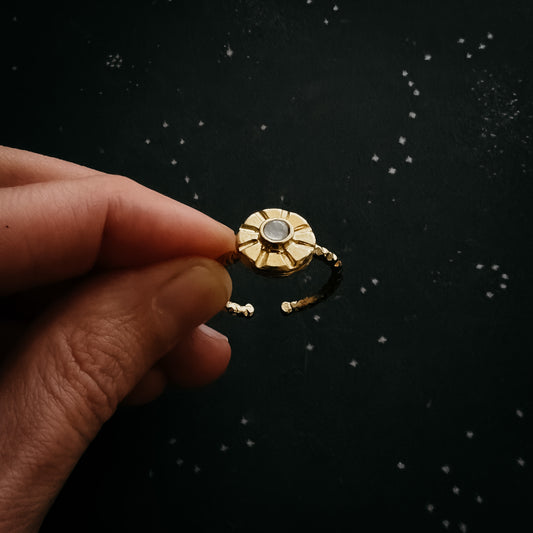 A hand holding a small gold ring with a circular sunburst design and a moonstone center, set against a dark starry background.