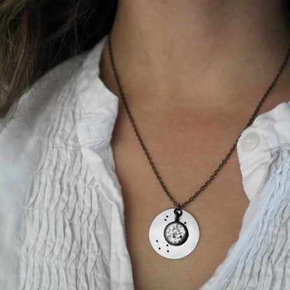 A close-up of a person wearing a silver pendant necklace with a circular disc charm featuring a central constellation cabochon, paired with a white textured blouse.