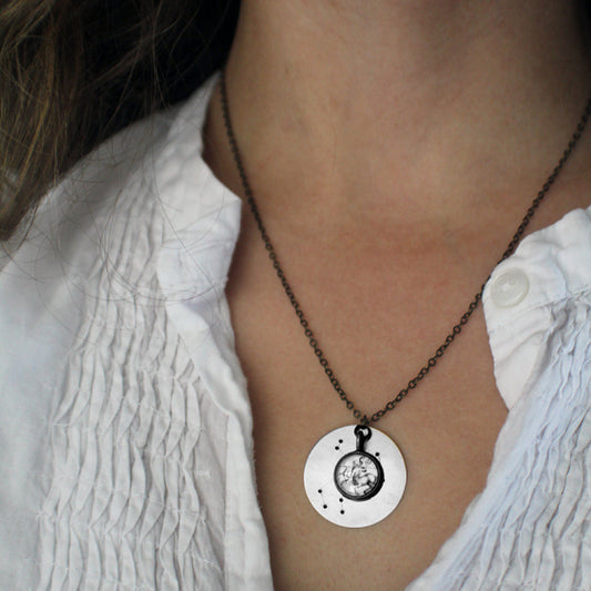 A close-up of a person wearing a silver pendant necklace with a circular disc charm featuring a central constellation cabochon, paired with a white textured blouse.