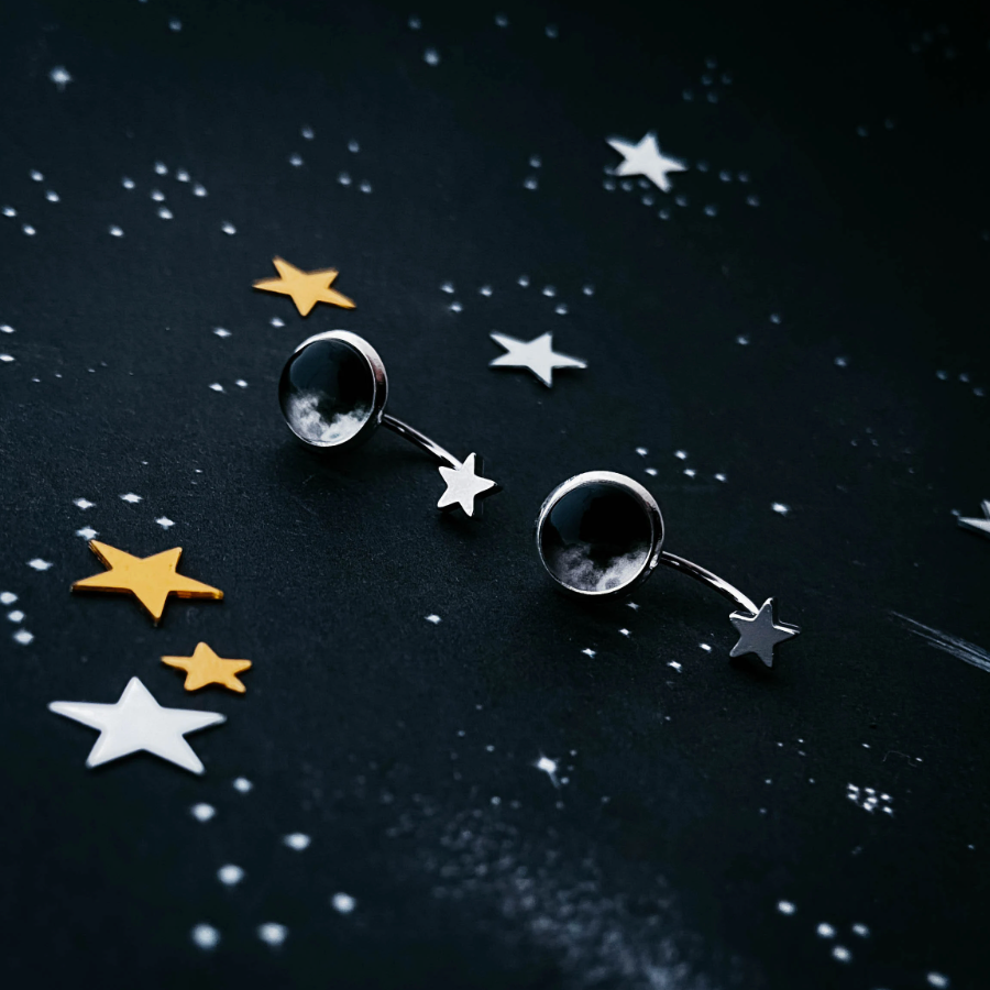A pair of silver-toned celestial stud earrings featuring a larger moon face pendant and a smaller dangling star, displayed on a dark starry background.