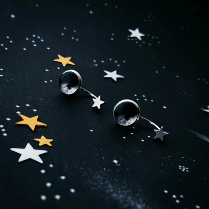 A pair of silver-toned celestial stud earrings featuring a larger moon face pendant and a smaller dangling star, displayed on a dark starry background.