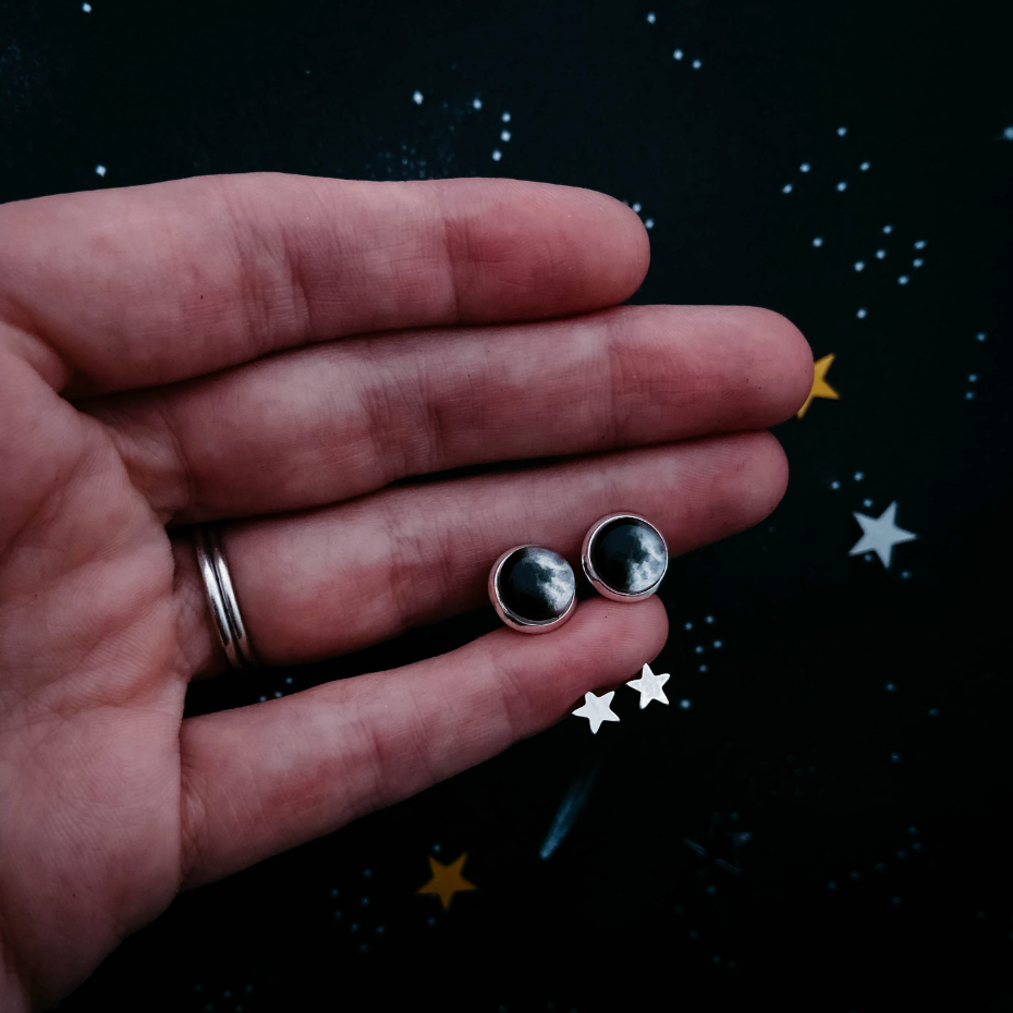 A hand holding a pair of silver moon phase earrings set against a dark starry background.