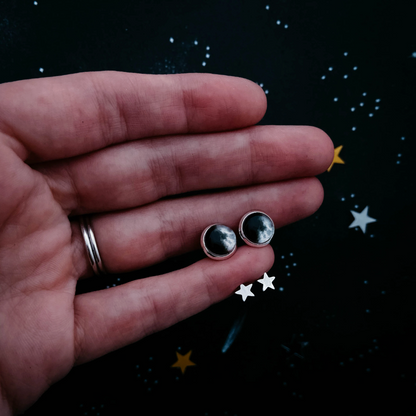 A hand holding a pair of silver moon phase earrings set against a dark starry background.