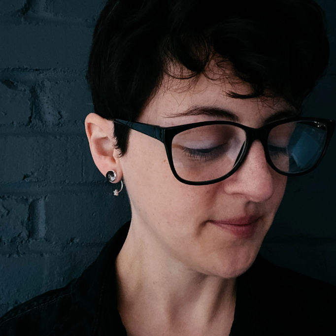 A close-up portrait of a person with short dark hair, wearing black-rimmed eyeglasses and a black top, standing against a dark blue-gray brick wall.