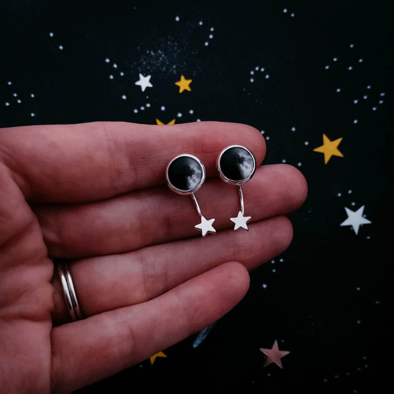 A hand holding a pair of silver moon and star earrings against a dark starry space background.