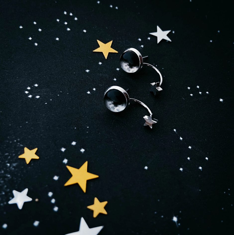 A pair of silver-toned earrings with moon phases and a hanging star, displayed on a dark textured background with scattered white and gold star confetti.