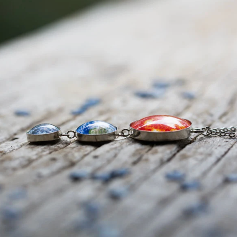 A close-up of a silver chain necklace with three small dome-shaped pendants resting on a weathered wooden surface. 