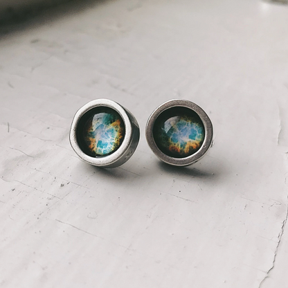 A pair of silver-toned stud earrings with round bezel settings containing a blue, green, and orange galaxy resin design, resting on a light gray textured surface.