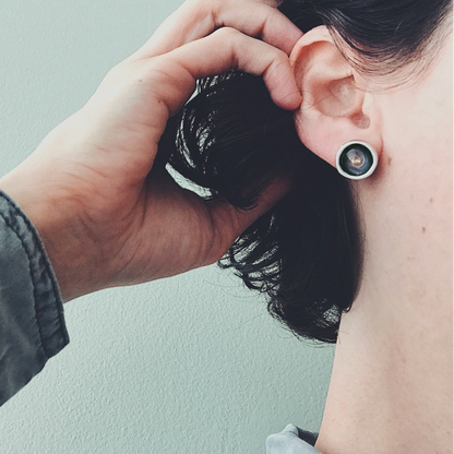 A close-up of a person's ear wearing a round silver-toned stud earring with a dark center, while another hand adjusts dark hair near the ear. The background is a plain light gray wall.