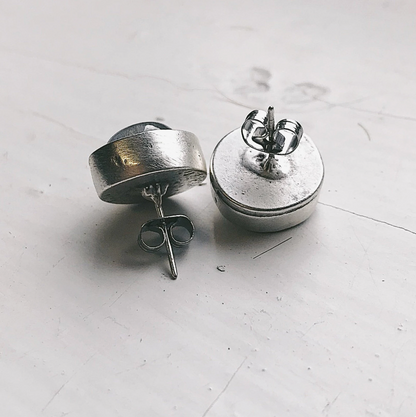 A pair of silver-toned stud earrings with a thick, chunky circular disc design, shown from both the back and side angles on a light gray surface.