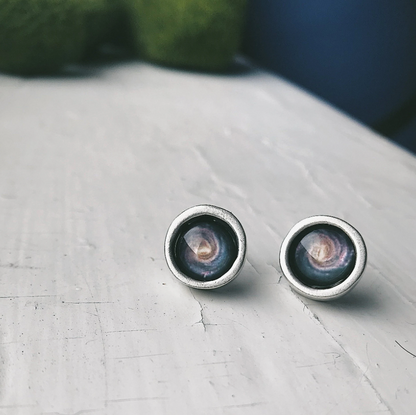 A pair of round stud earrings with silver-toned metal rims and a milky way swirl image, resting on a light-colored wooden surface.