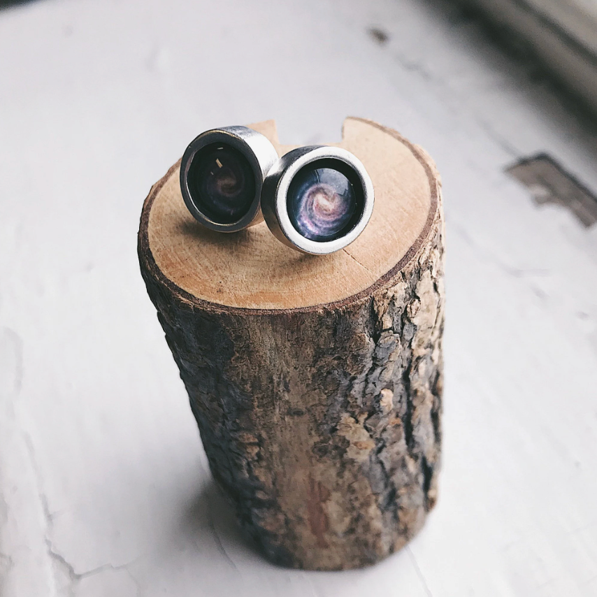 A pair of silver-toned stud earrings with detailed milky way image, displayed on a rustic wood log slice against a light textured background.