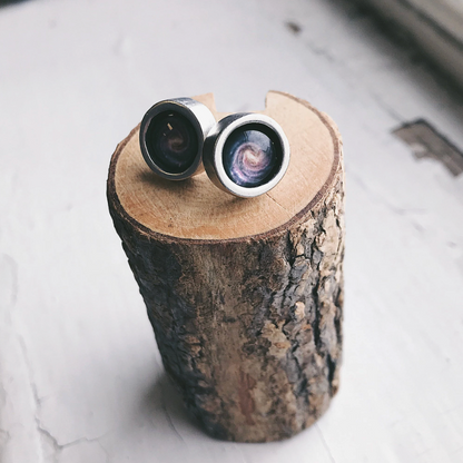 A pair of silver-toned stud earrings with detailed milky way image, displayed on a rustic wood log slice against a light textured background.