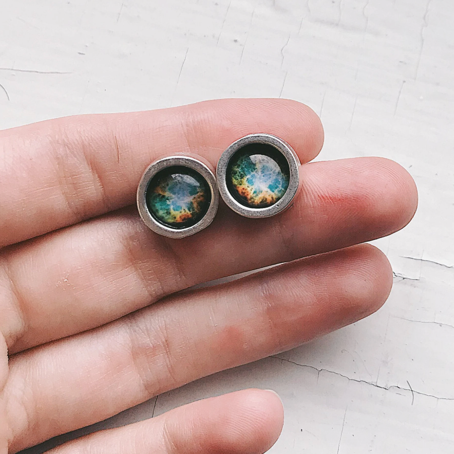 A pair of round stud earrings with silver-toned metal rims and colorful galaxy-like centers, shown resting on a person's fingers against a light textured background.