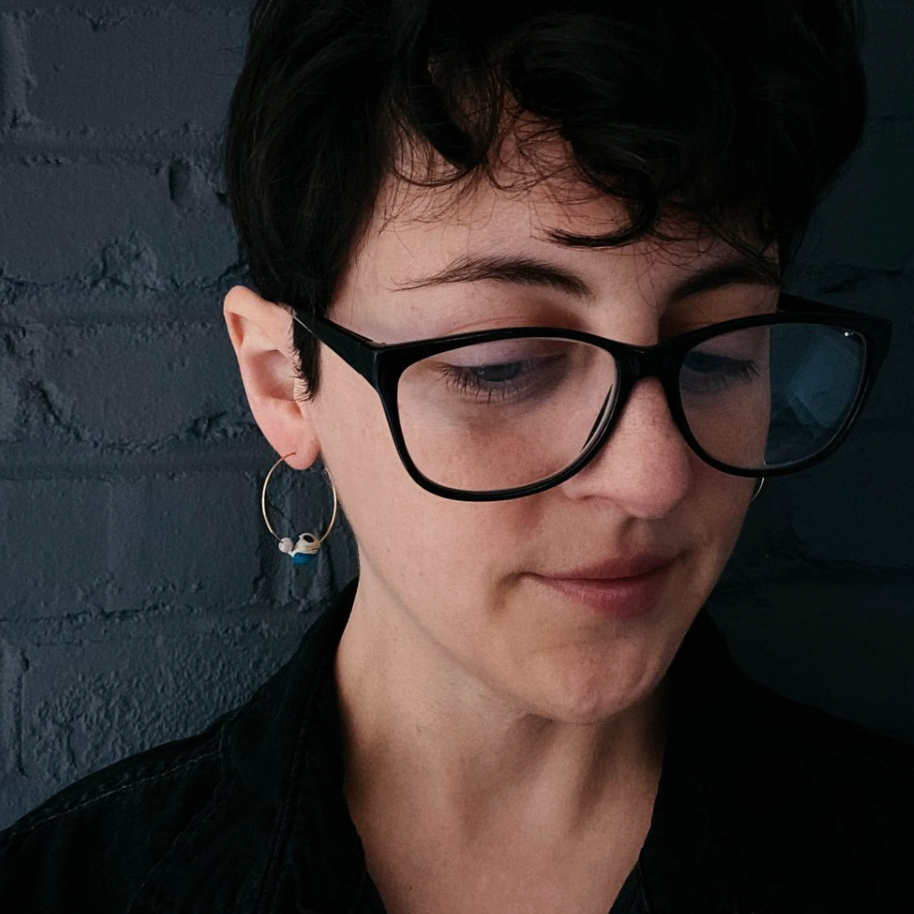 A close-up portrait of a person with short dark hair wearing black glasses and a small silver hoop earring, set against a dark textured brick wall.