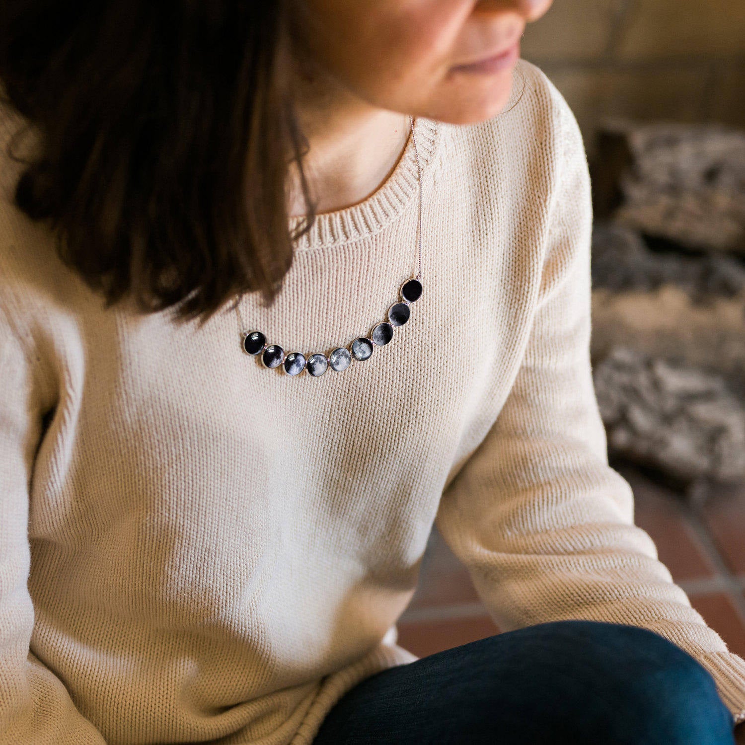 Person wearing a beige sweater with a moon phase necklace, sitting indoors.