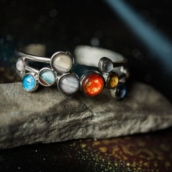 A silver-toned ring featuring multiple round planet cabochons, displayed on a textured gray stone against a dark, starry background.