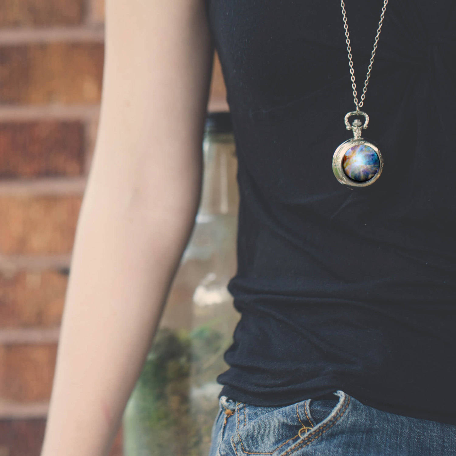 A person wearing a silver-toned chain necklace with a round pendant featuring a colorful galaxy nebula design, styled with a black t-shirt and blue jeans.