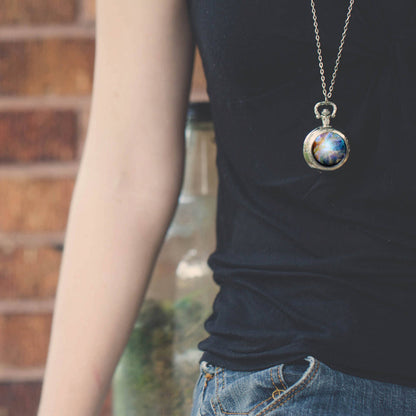 A person wearing a silver-toned chain necklace with a round pendant featuring a colorful galaxy nebula design, styled with a black t-shirt and blue jeans.