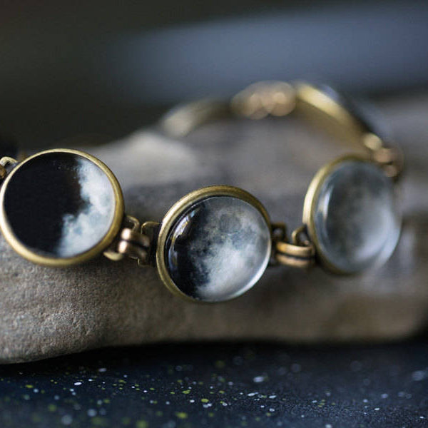 A close-up of a bracelet with moon phase cabochons set in bronze-tone metal, resting on a gray stone surface.