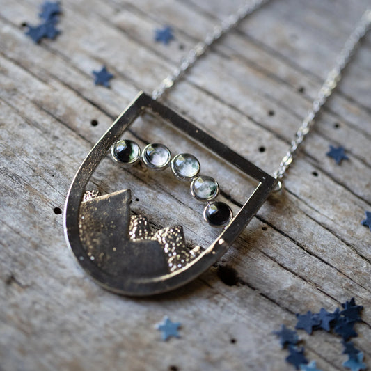 A silver shield like pendant with a mountain scene and 5 moon phases overhead, resting on a wooden background with blue star confetti. 