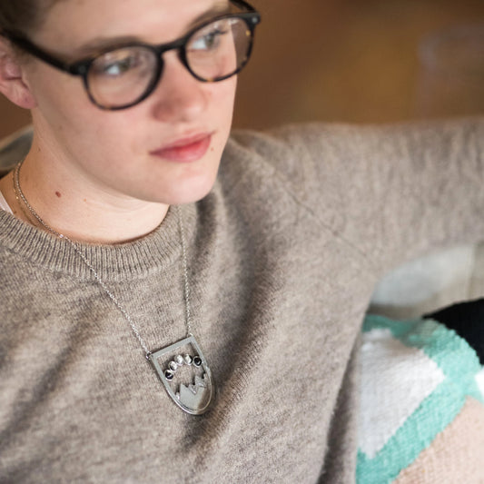 Woman with glasses and a sweater wearing a moon phase mountain landscape necklace. 