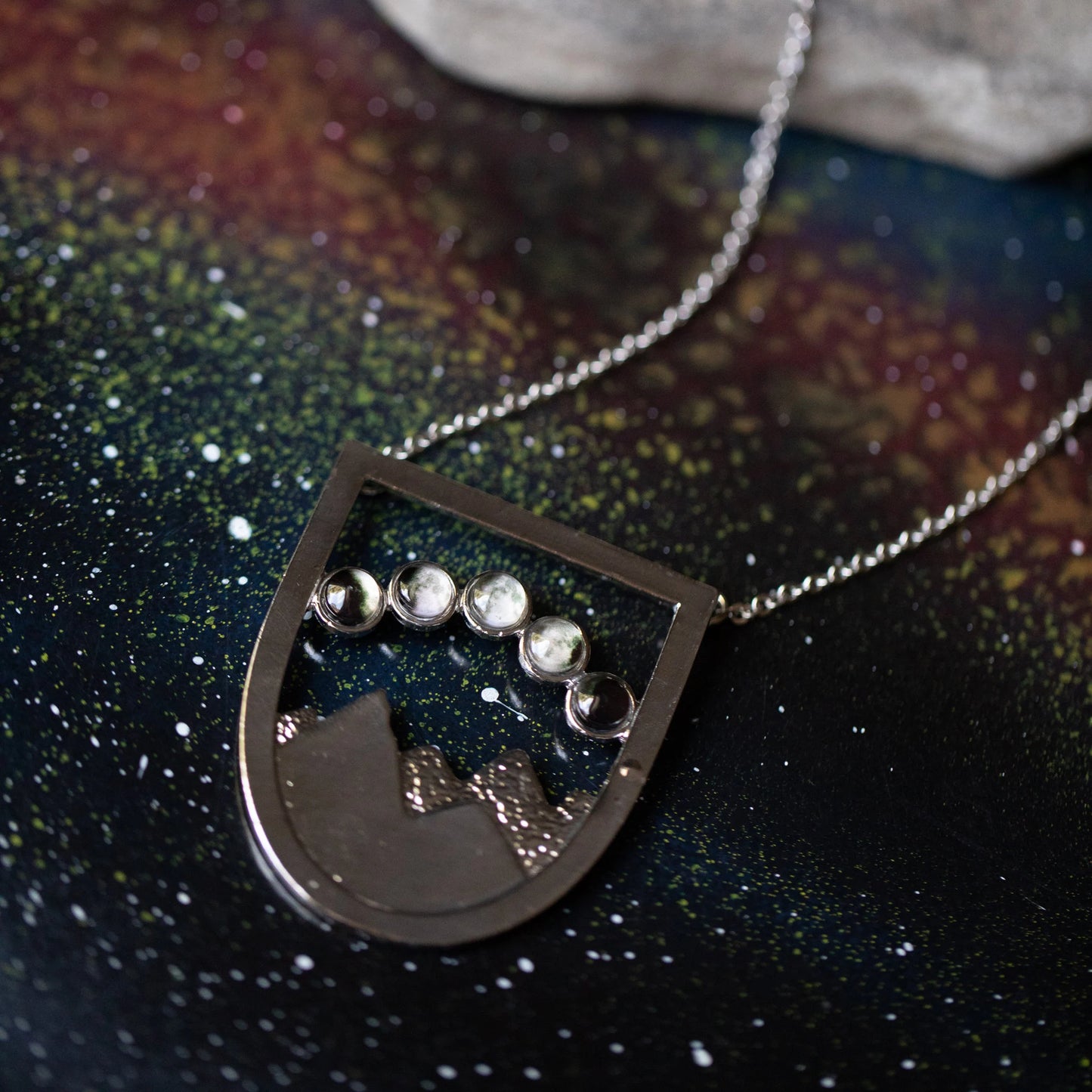 A silver shield like pendant with a mountain scene and 5 moon phases overhead, resting on a dark galaxy like background. 