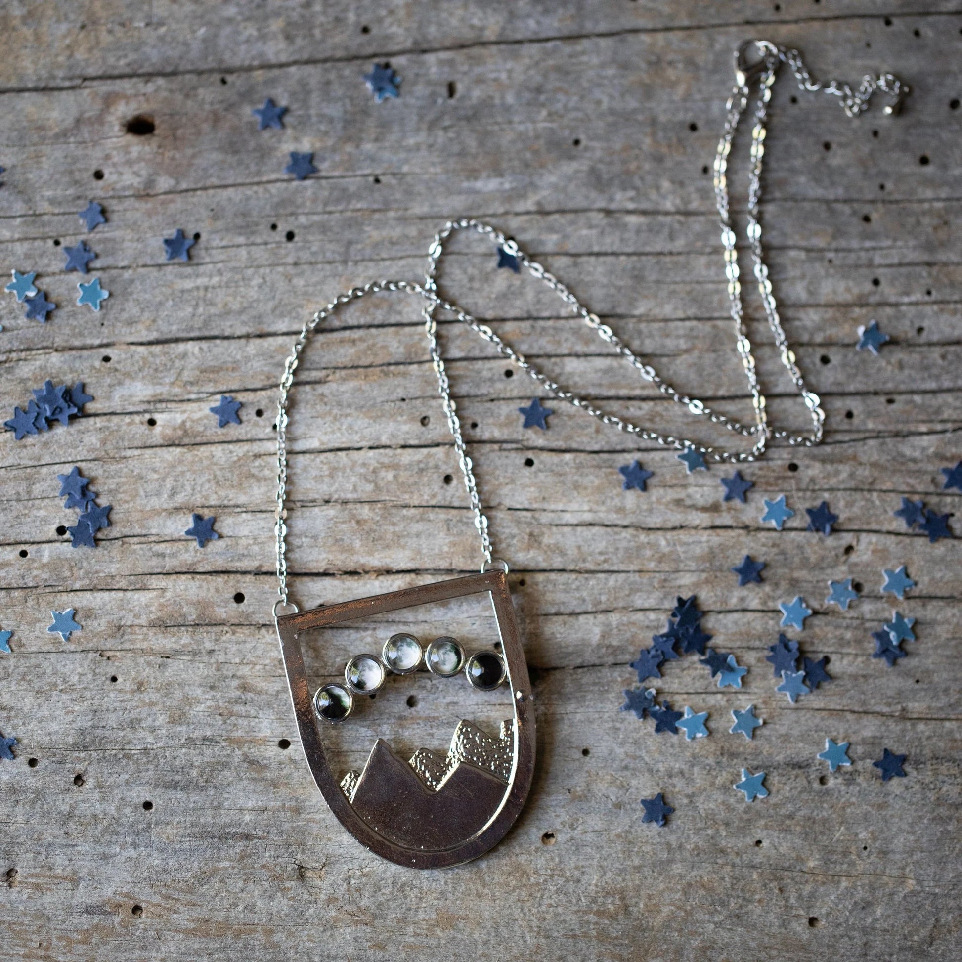 A silver shield like pendant with a mountain scene and 5 moon phases overhead, resting on a wooden background with blue star confetti. 